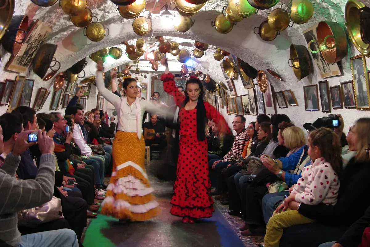 Two flamenco dancers in a red polka-dot dress and a yellow ruffled skirt on a narrow floor in a whitewashed cave hung with brass pans, with spectators seated along both walls